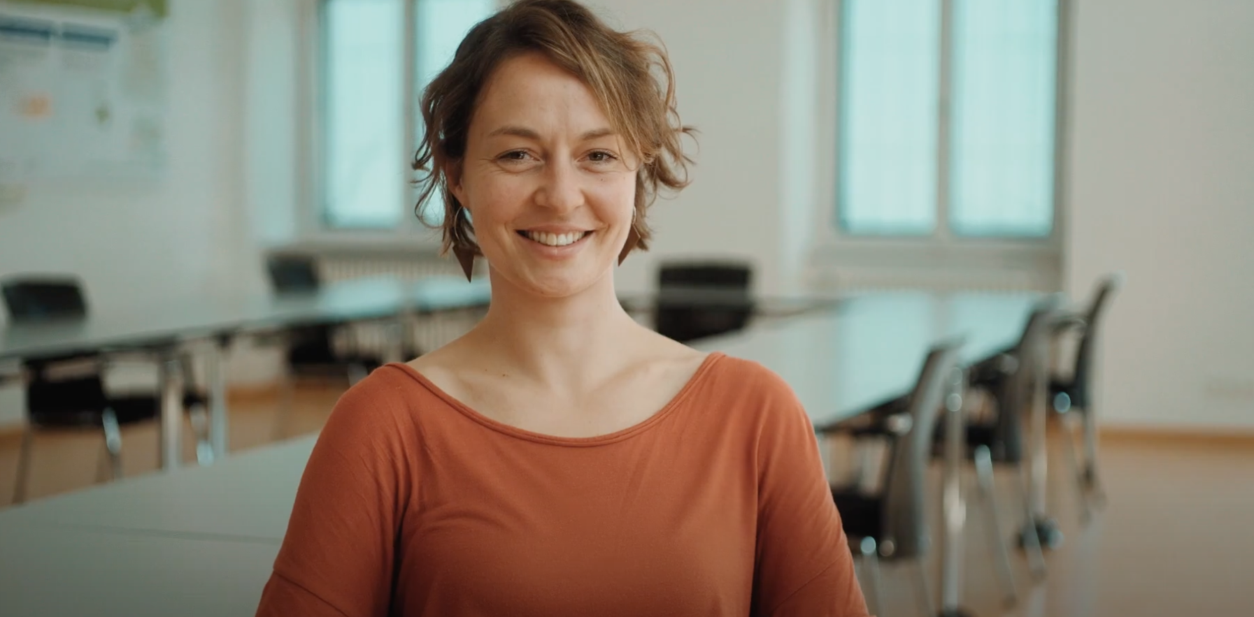 Frau sitzt in einem Konferenzraum. Stühle und Tisch im Hintergrund.
