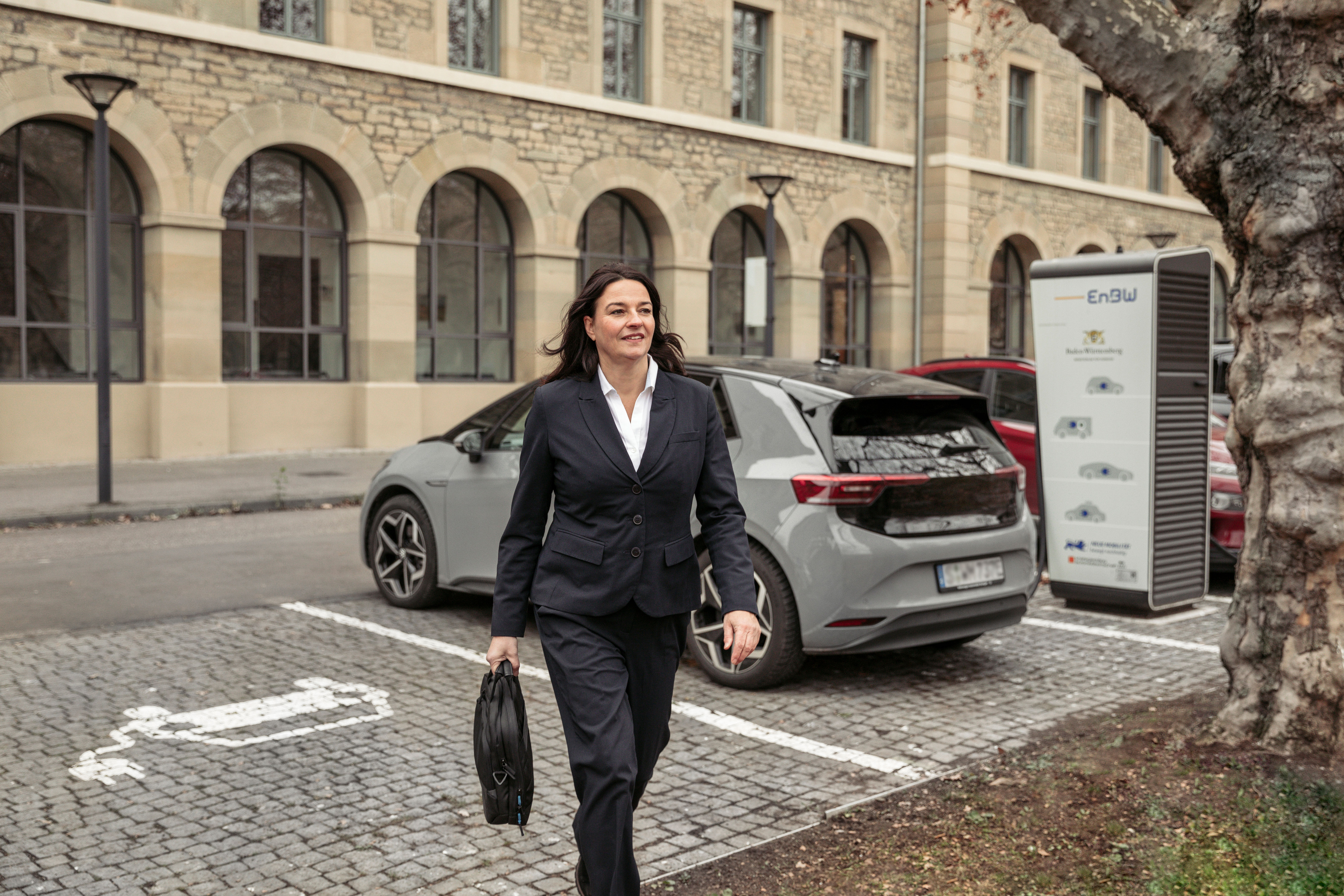 Geschäftsfrau mit Aktenkoffer geht von einem grauen E-Auto weg, das an einer Ladesäule lädt.