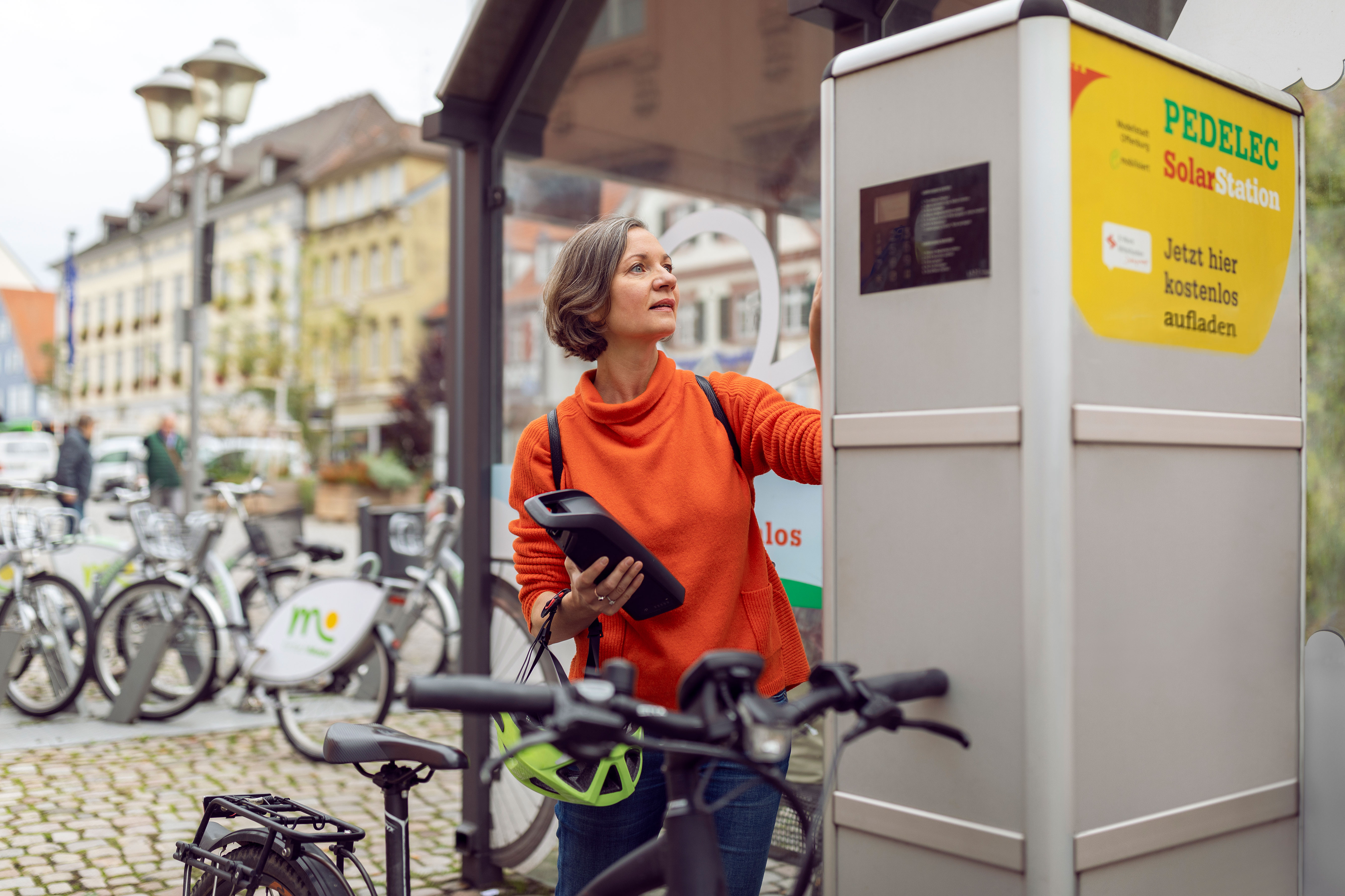 Frau lädt Pedelec-Akku an einer Solar-Ladestation in der Stadt, Fahrräder im Hintergrund.