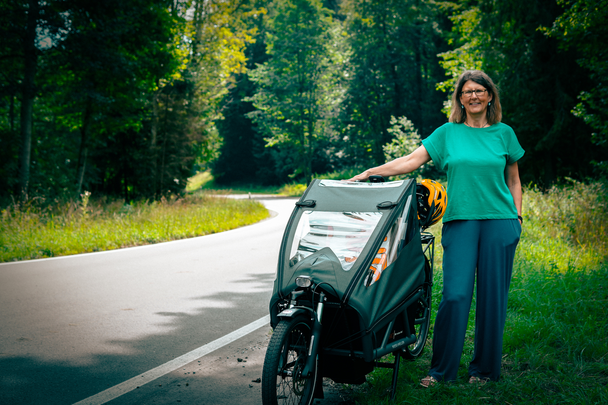 Eine Frau steht mit ihrem Fahrrad am Straßenrand einer Landstraße im Wald.