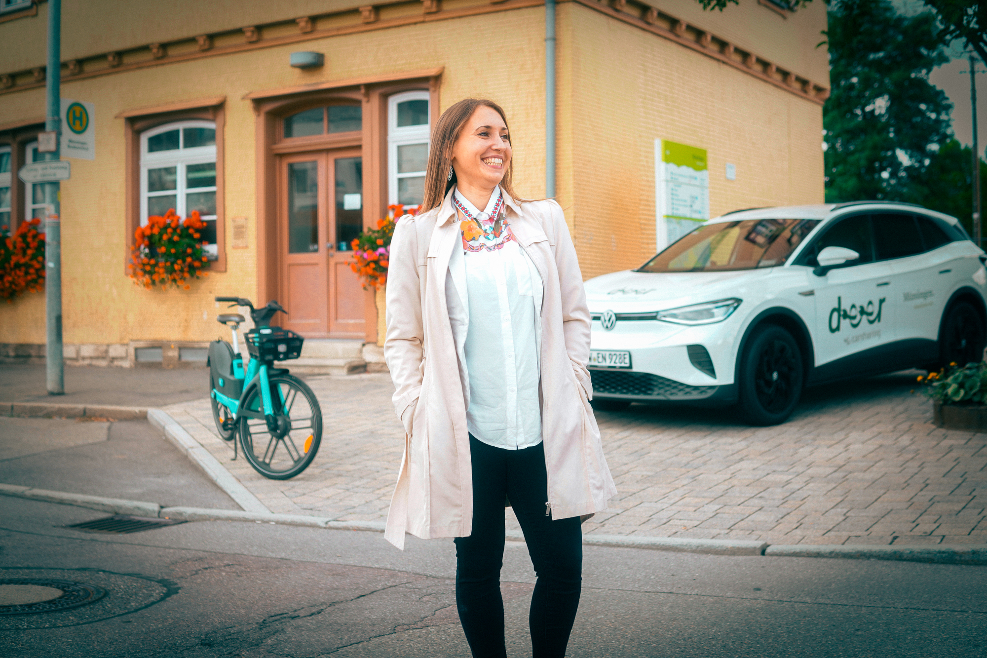Eine Frau steht lachend vor einem Gebäude, im Hintergrund steht ein Auto und ein Fahrrad.