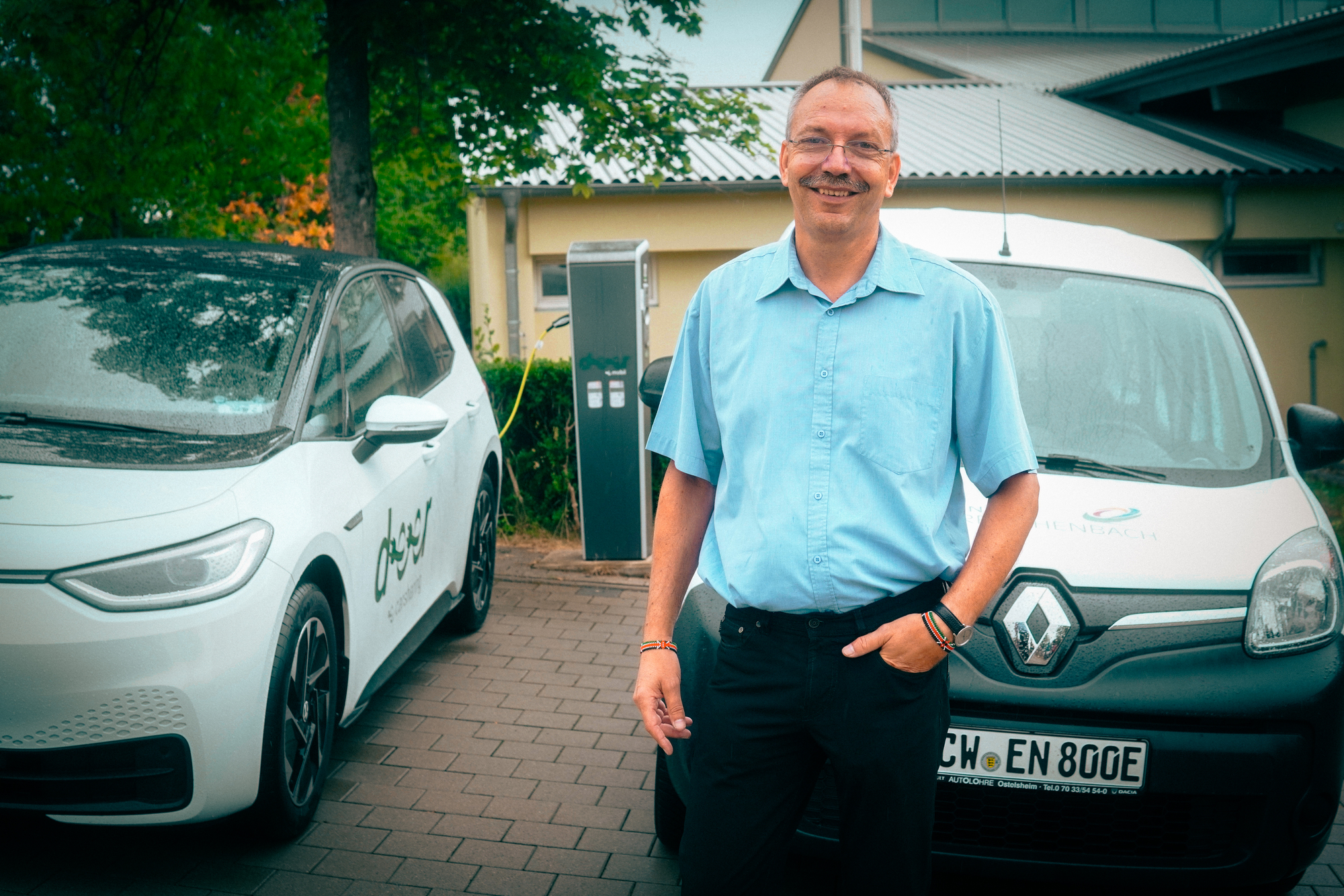 Ein Mann im blauen Hemd steht vor zwei Elektroautos welche an einer Ladesäule geladen werden.