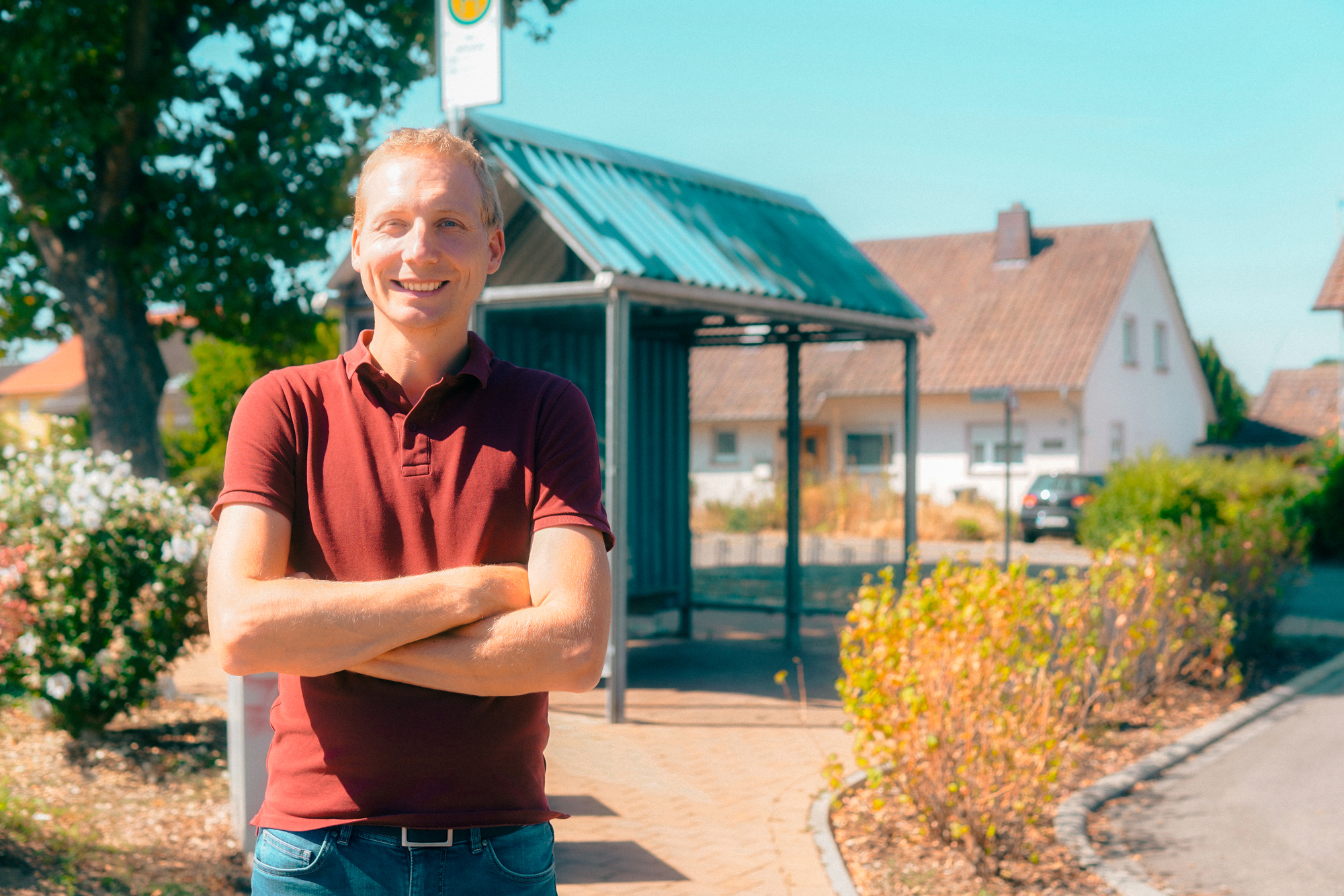 Ein lachender Mann der im Sonnenlicht vor einer Bushaltestelle steht.