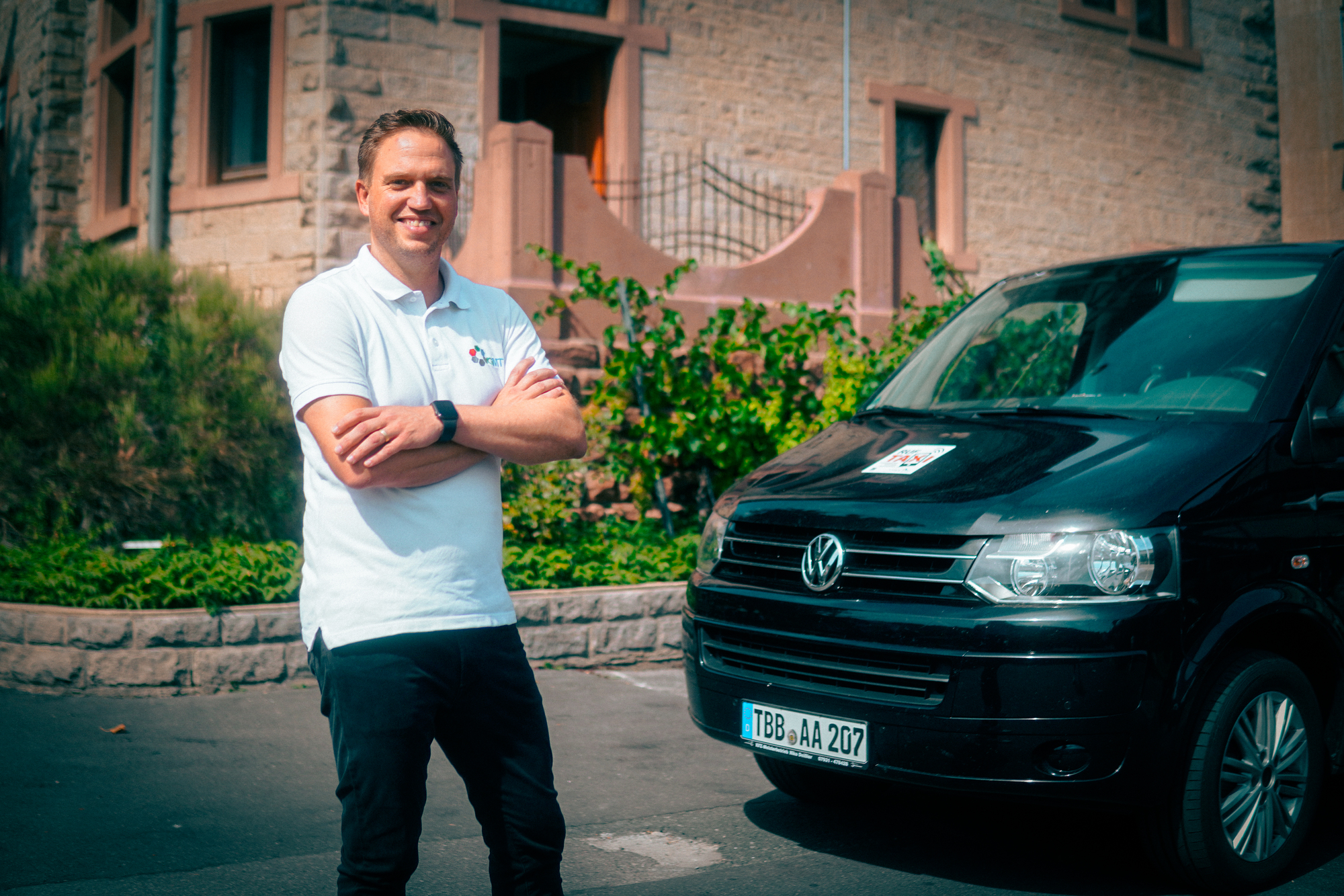 Ein Mann steht mit verschränkten Armen vor einem Gebäude, an der Seite ist ein schwarzes Auto zu sehen.