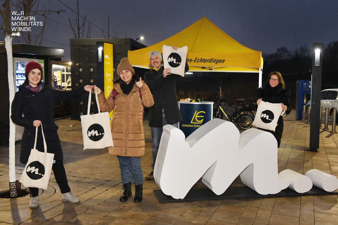 4 Personen die in der Dunkelheit posieren vor einem Pavillon und strecken Jutebeutel lächelnd in die Luft.