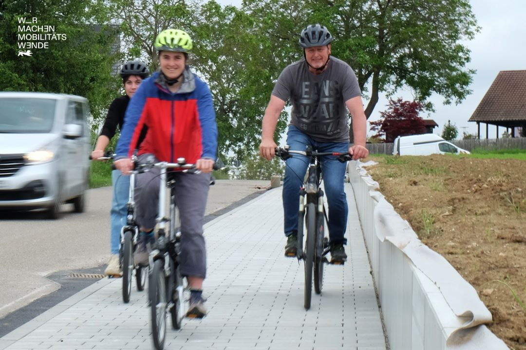 Drei Personen auf unterschiedlichen Fahrrädern die neben der Straße fahren. Ein Auto im Hintergrund.