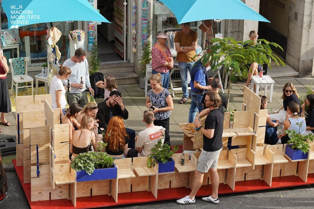 Eine große Gruppe Menschen die auf Sitzmöglichkeiten aus Holz vor einem Haus sitzen und sich unterhalten.
